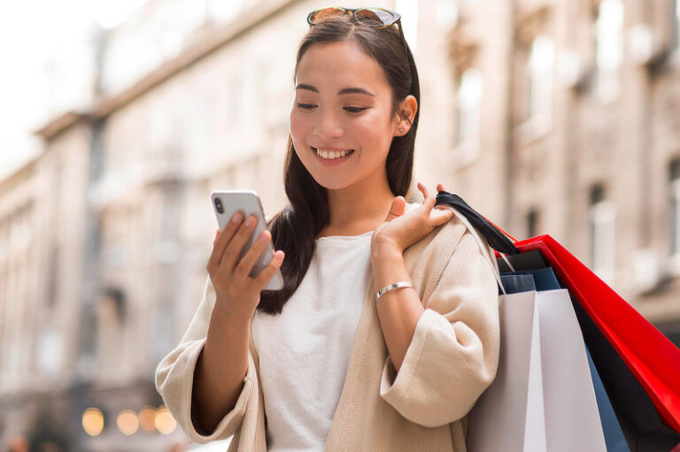 Woman looking at smartphone outdoors while holding shopping bags.